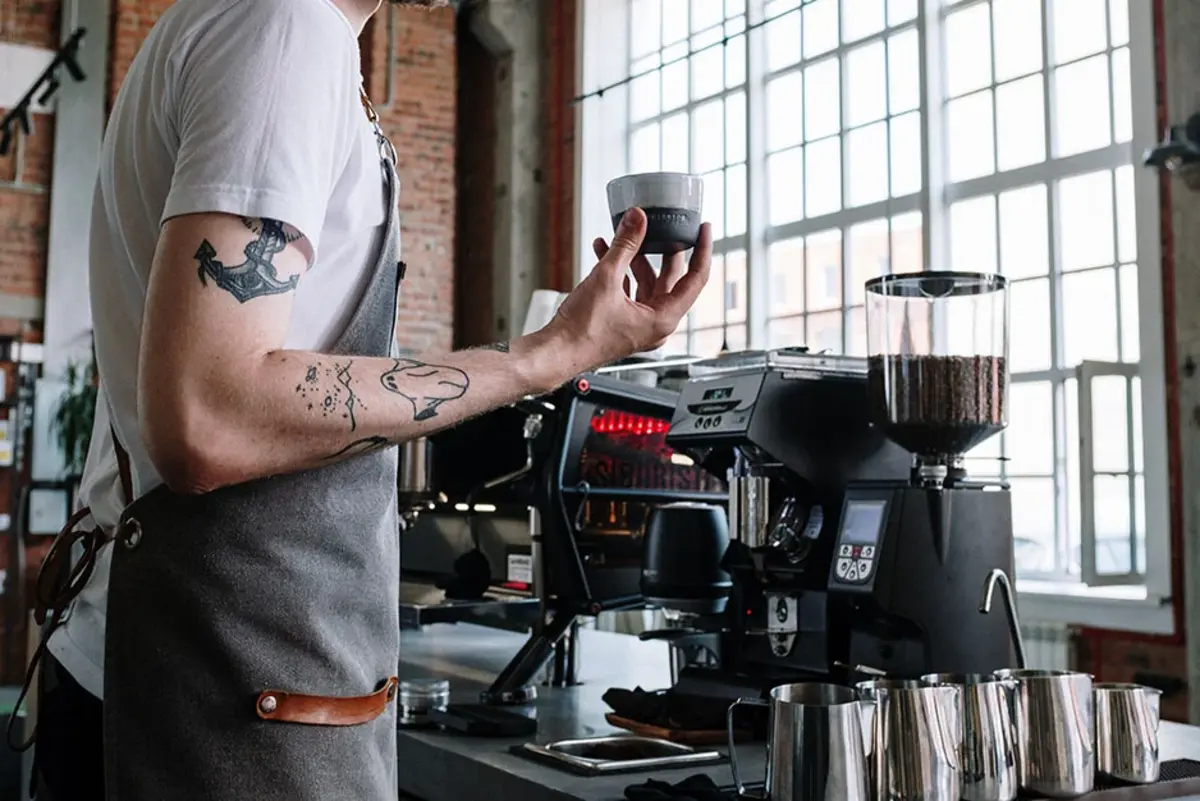 Barista holding a cup beside a two-group commercial espresso machine and bean grinder